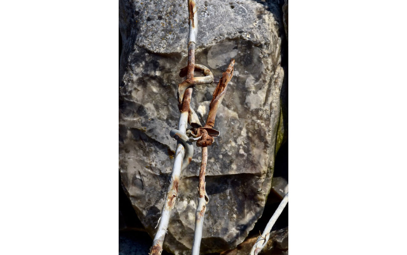 Le visage carré taillé à la serpe est comme balafré par le grillage qui lui traverse le visage. Une pointe du fil de fer du gabion menace de lui percer l'oeil. L'enfer du gabion 15-La pointe rouillée-Les pierres du chemin-Photo J Lamy 2019.