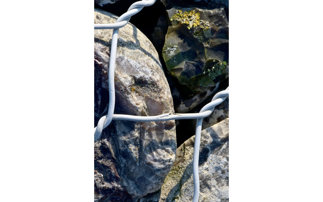 Appuyé contre le grillage, un visage triste. en second plan, dans l'ombre un visage anguleux presque menaçant. L'enfer du gabion 12-Les deux frères-Les pierres du chemin-Photo J Lamy 2019.